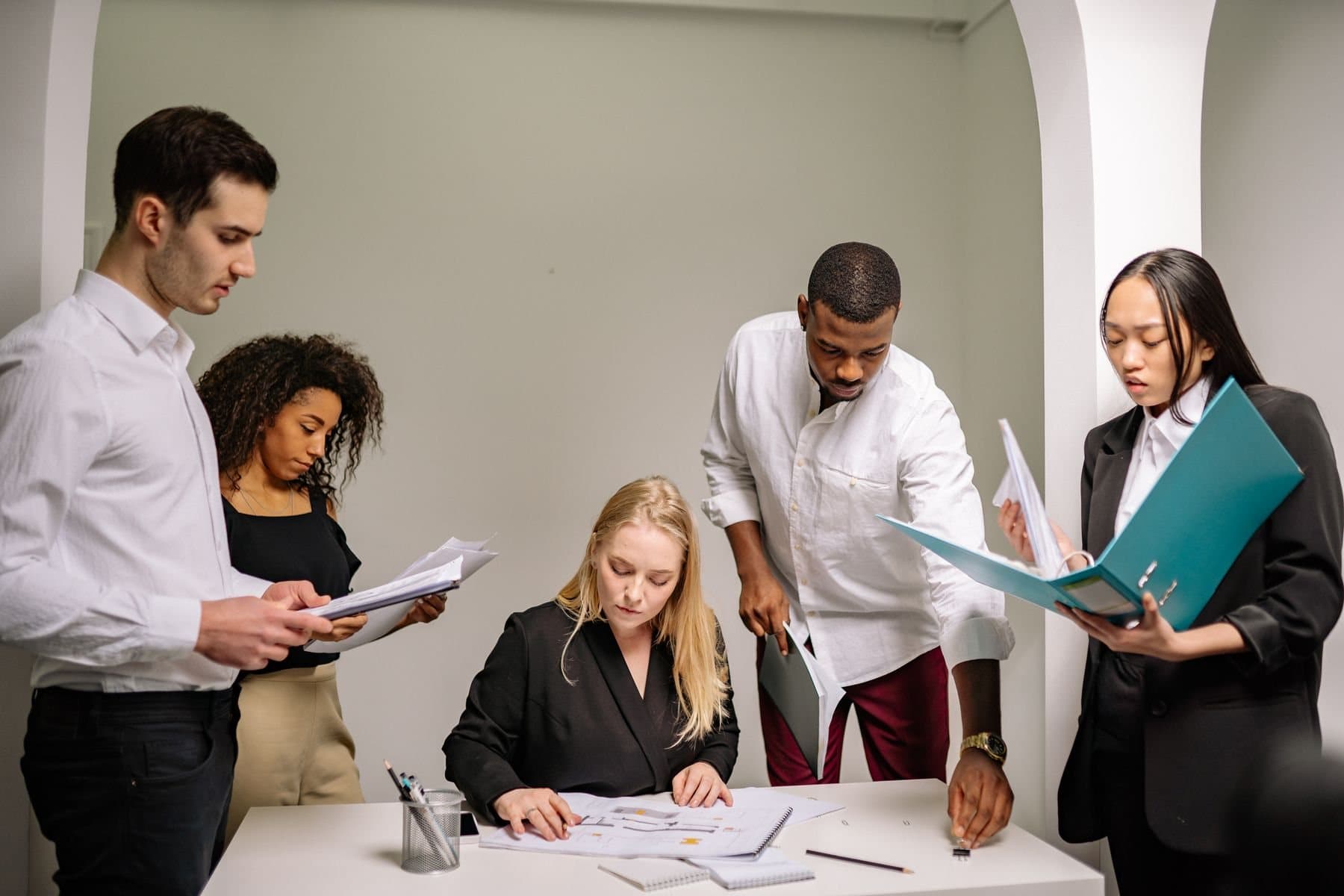 Five colleagues around a desk, several holding folders of printed work